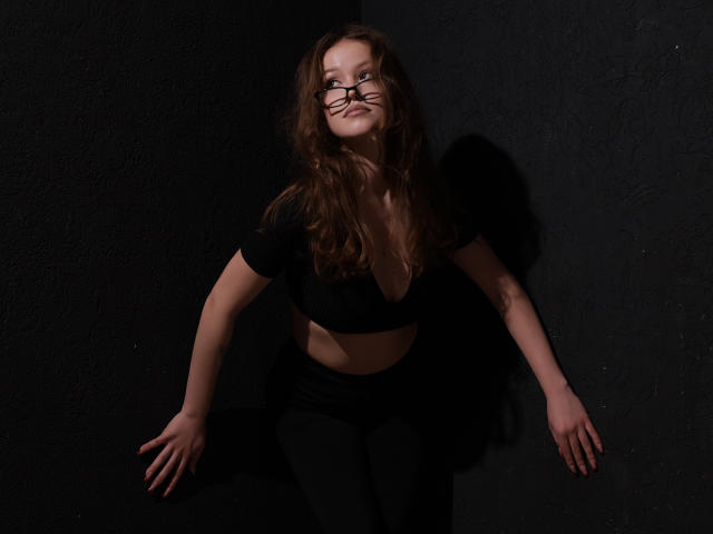 BeatriceKeat with wavy brown hair, black crop top, and black pants, leaning against a dark wall, biting glasses, arms out, smiling slightly, shadowed, mid-shot, looking up.