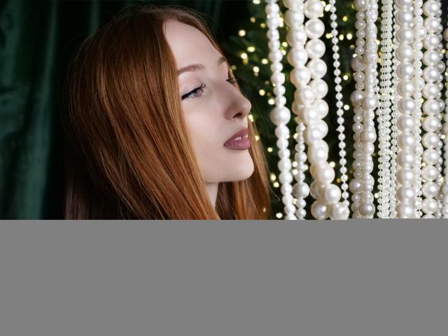 TinaSerenade has long red hair, pale skin, and is looking to the side with her eyes closed. She's wearing a green ribbon with white pearl chains in front.