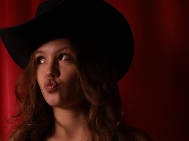 BeatriceKeat has wavy brown hair, a black hat, and pouty lips, looking to the side with a slight smile, against a red background. Her tits are not visible.