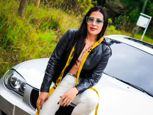 JessyLuxs with long black hair, wearing a black leather jacket, yellow straps, and white pants, sitting on a white BMW, wearing green sunglasses, showing small tits, and a necklace.