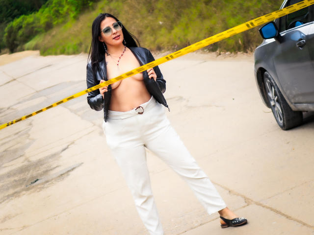 JessyLuxs with black leather jacket open, showing her tits, wearing white pants, black flats, yellow tape in front, standing on concrete, next to a gray car.