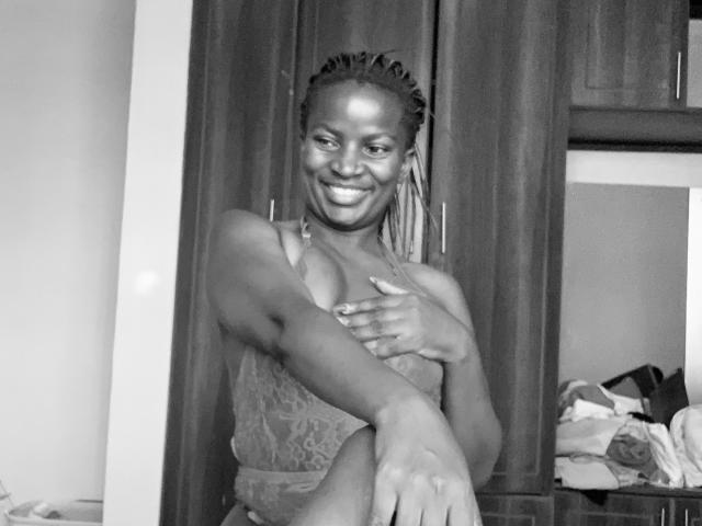 JennyNorah with dark skin, smiling, wearing a lace top, covering her tits with one hand, sitting in a room, wood cabinets in the background, looking happy and relaxed.