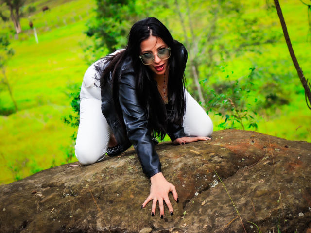 JessyLuxs with long black hair, wearing glasses and a black leather jacket, is on all fours on a rock, looking back with a red-lipsticked, slightly open mouth.