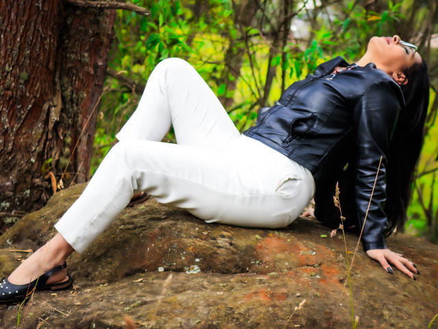 JessyLuxs is laying back on a rock, wearing white pants and a black leather jacket, head tilted up, black flats, and long black hair, in a forest.