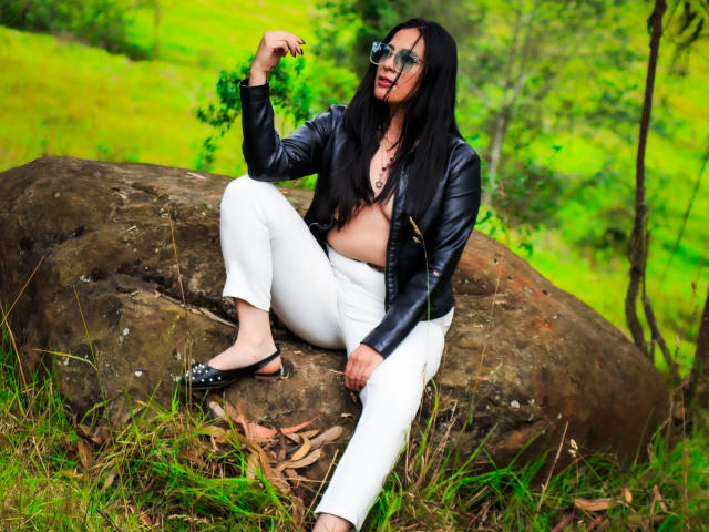 JessyLuxs with black hair, leather jacket, white pants, and black flats, sitting on a rock, showing one breast, wearing glasses, holding a leaf, in a green forest.