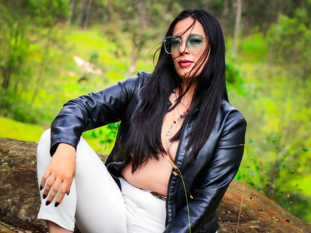 JessyLuxs with black leather jacket, white pants, and open shirt, exposing her tits, sitting on a rock, wearing green sunglasses, long black hair, and red lipstick, in a forest.