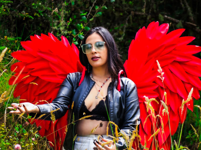 JessyLuxs with red feather wings, black lace bra, leather jacket, and green sunglasses, standing in wild grass, looking confident with hands out. Boobs visible, red wings are huge.