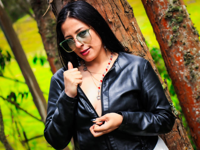 JessyLuxs with black leather jacket, green glasses, and red bead necklace, giving a thumbs-up, showing a hint of her tits, standing against a tree.