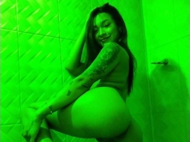 SashaHans with tattoos, smiling, nude, green-lit, leaning against a wall, showing her big ass, long brown hair, and right arm tattoo, holding her hair with her left hand.