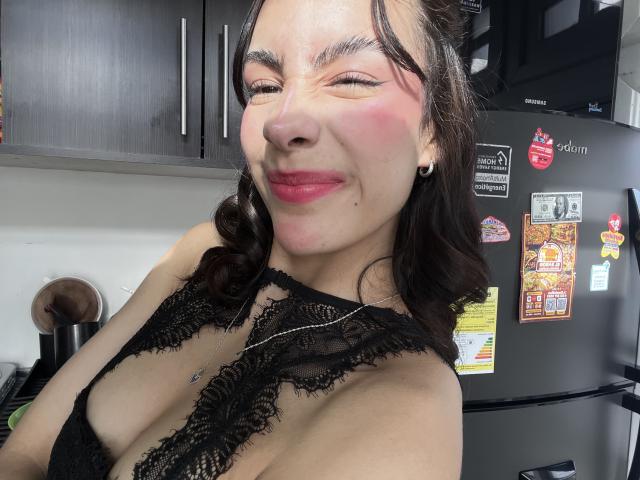 AitanaJohns with wavy brown hair, closed eyes, red lipstick, and black lace top, showing her tits and smiling. She's in a kitchen with a dark fridge.