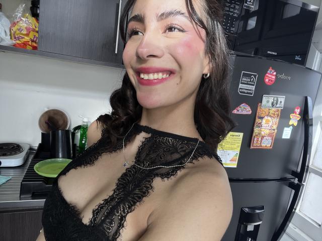 AitanaJohns, smiling with pink blush on cheeks, wearing black lace bralette, showing her tits, in kitchen with dark fridge and magnets, has curly brown hair, red lipstick.