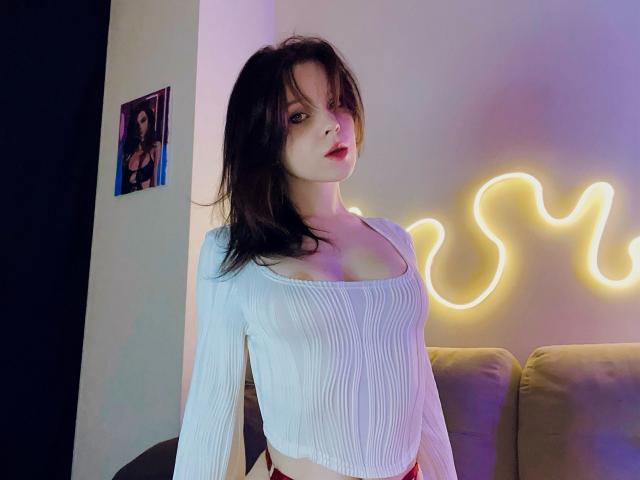 lkPleasure with pale skin and dark brown hair, wearing a white, ribbed, long-sleeve top, showing her small tits, standing in front of neon yellow wall art.