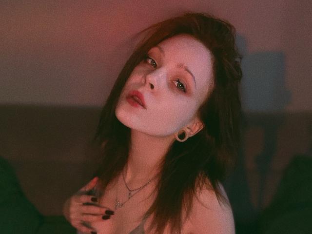 LK Pleasure with pale skin, dark brown hair, and red lipstick, wearing a necklace and earring, looking up with a slightly open mouth, hand on chest, in dim light.