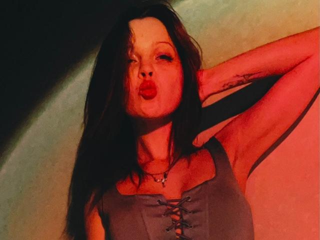 lkPleasure with pouty red lips, dark wavy hair, gray lace-up top, tattooed arm, and necklace, leaning against wall in red and green light.