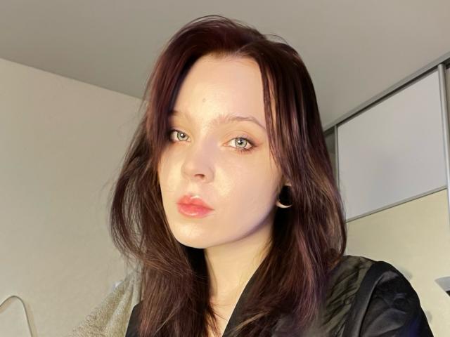 lkPleasure has pale skin, green eyes, and red lipstick. She has dark brown, slightly messy hair and is wearing a black shirt with black ear cuffs.