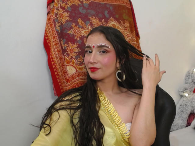 JalimaMona, com cabelos longos e pretos, batom vermelho, blush rosa, joias douradas e um sari amarelo, tocando o cabelo, usando uma peça decorativa na testa, sentada em uma cadeira preta..