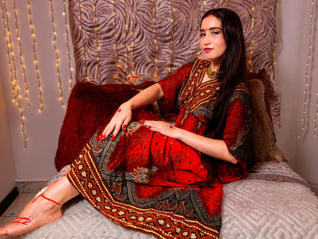 JalimaMona with dark hair, red dress, and ornate patterns, sitting on a bed, showing off her bare ass with red string on her foot, and red bangle.