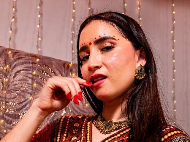 JalimaMona with red dot makeup on forehead, wearing red lipstick and earrings, touching her red-painted fingernails, in a brown patterned top, with a gold necklace.