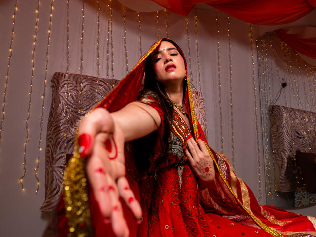 JalimaMona with red and gold traditional dress, reaching out with red-painted fingers, veil over head, jewelry, and henna on hands, looking seductively at camera, medium shot.