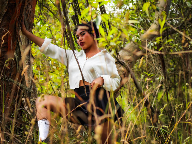 SophieSonyas with medium tits, black ass, wearing a white crop top, black shorts, and white socks, standing against a tree in a forest, looking at the camera.