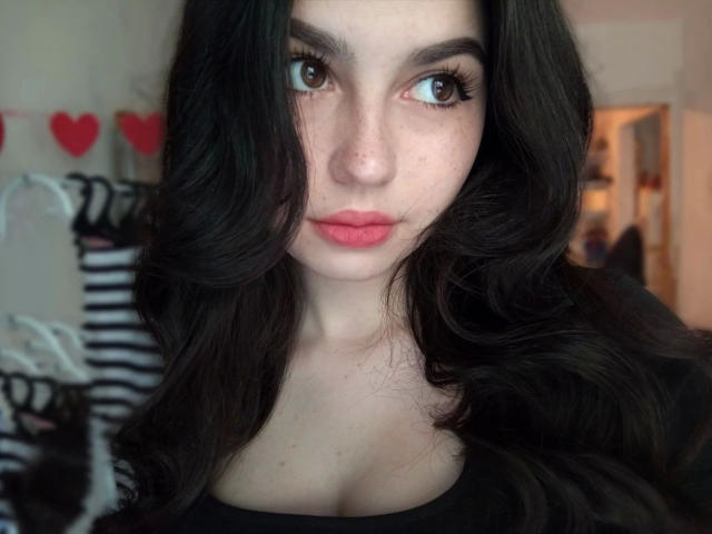 Close-up of KisaModel-hot with long, wavy black hair and fair skin. She has large brown eyes with long lashes, pink lips, and a slight freckle pattern on her cheeks. She's wearing a black top. Background is blurred with red heart decorations and a black and white striped garment. The image has a soft focus, emphasizing her facial features and hair texture.