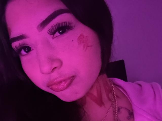 NovaTooNaughty's face, purple-lit, with thick eyebrows, long eyelashes, glossy lips, tattoo near right eye, necklace, and tattoo on neck. Her expression is slightly pouty.