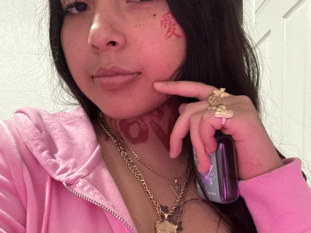 NovaTooNaughty with a pink hoodie, holding a phone, has tattoos on her neck and cheek, and wears gold chains and rings, with pink nails, in a selfie.