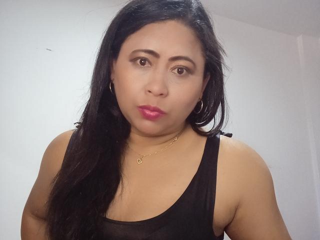MiaSalomeChaude has medium brown skin, long black hair, pink lipstick, hoop earrings, a black tank top, and a gold necklace, looking directly at the camera with a serious expression.