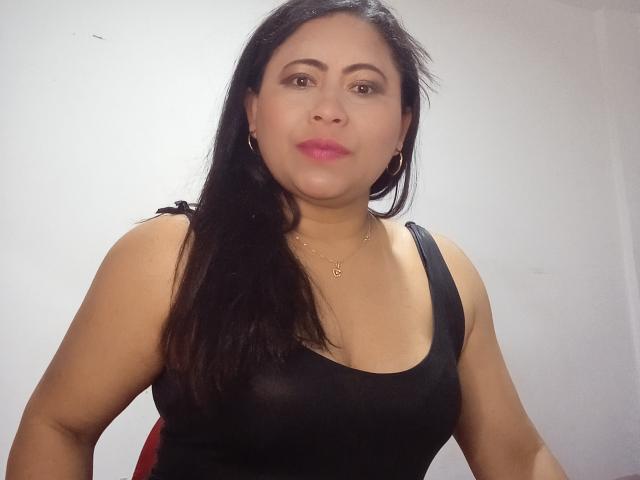 MiaSalomeChaude with dark long hair, pink lipstick, black tank top showing her tits, gold necklace, and hoop earrings, standing against a white wall.