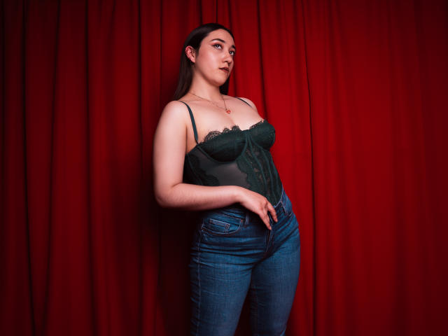 SwettieSammy draagt een donkergroene body met veters, waarbij haar borsten zichtbaar zijn, en strakke blauwe jeans. Ze staat voor een rode gordijn, kijkt naar de zijkant en heeft haar hand op haar buik. Ze is schattig, met een lichte huidskleur..