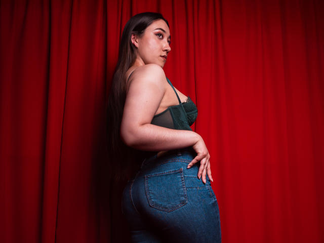 SwettieSammy, avec ses longs cheveux bruns, son soutien-gorge vert et son jean bleu, montrant ses courbes et sa poitrine, debout devant des rideaux rouges, regardant par-dessus son épaule..