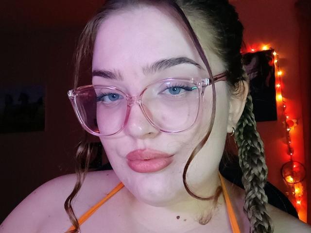 TeasingXDora with puffy pink lips, wearing clear glasses, orange halter top, and braided brown hair, has blue eyes and fair skin, showing a slight pout.