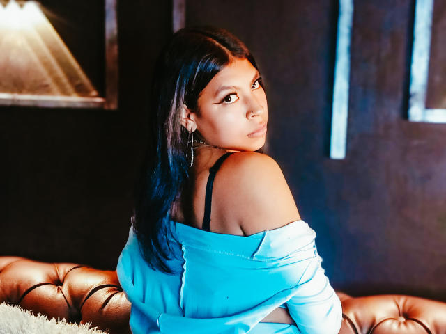 SashaLia with dark hair, looking over her shoulder, wearing a blue off-shoulder top, black bra straps visible, sitting on a brown leather couch, medium tits.
