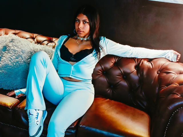 SashaLia is sitting on a brown leather couch, wearing a blue zip-up top and matching pants, showing black lace bra, with a fluffy gray pillow on her left.