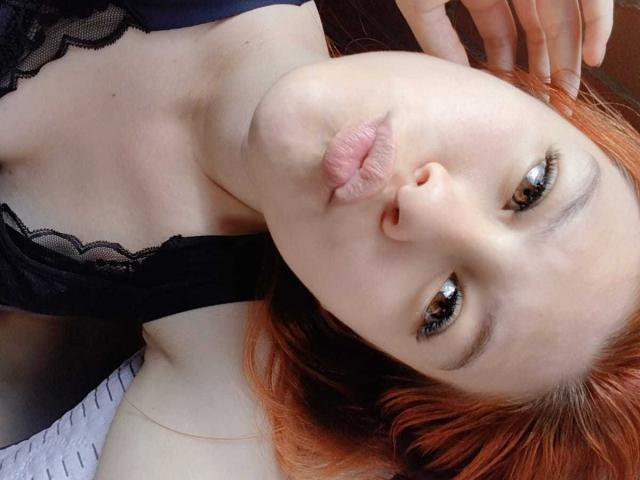 TaylorCarpert with pouty pink lips, red hair, and black lace top, looking up with half-closed brown eyes, her hand near her head, showing a cute, sexy face.
