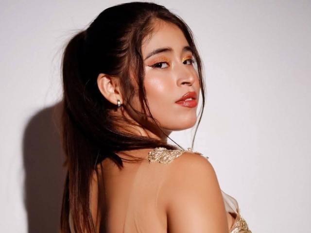 AlanaKlass with long dark ponytail, looking back, wearing sheer top with gold floral details, red lipstick, and winged eyeliner, showing her glossy, shiny ass.