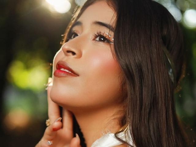 AlanaKlass has glossy brown hair, glowing skin, and red lipstick. Her hand touches her chin, looking up with sparkling eyes. Sunlight flares in the background.