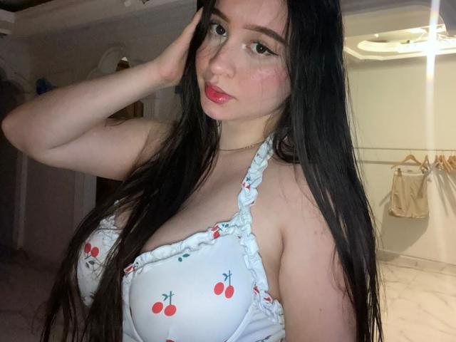 SophieHale with long black hair, wearing a white cherry-patterned halter top, exposing her large tits, hand on her head, pink lipstick, and a cute face in a dimly lit room.