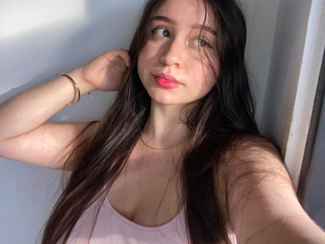 SophieHale with long brown hair, pink tank top, pink lips, and bracelet, looking at camera with hand in hair, pale skin, slight blush, and soft light.
