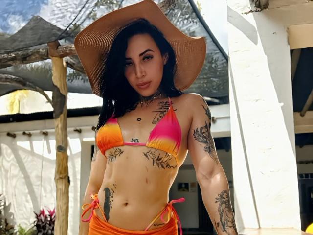 YeiSela with tan skin, black hair, and tattoos, wearing an orange-yellow bikini, tan cowboy hat, showing her boobs and ass, standing outside in bright sunlight.