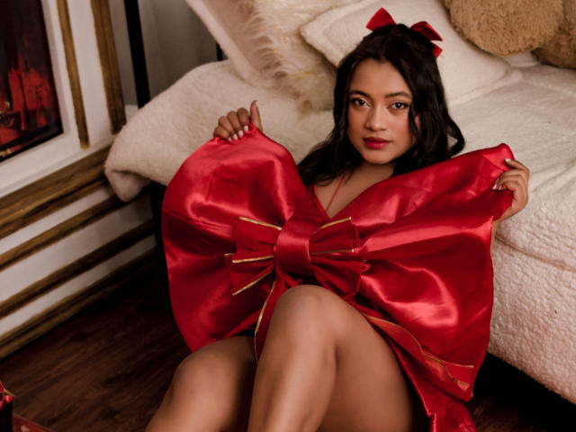 Sarita, charmante, porte une robe en satin rouge avec un nœud. Elle la tient ouverte pour révéler ses seins et ses parties intimes, assise sur un plancher en bois. Elle a les cheveux longs et ondulés, des lèvres rouges et un lit blanc en arrière-plan..