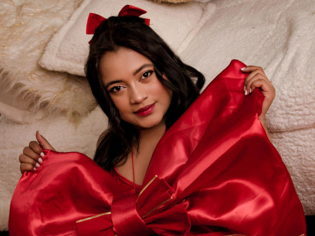 Sarita, com cabelos castanho-escuros, um laço vermelho e um vestido de cetim vermelho, está segurando-o aberto para mostrar seus pequenos seios adoráveis. Ela está sorrindo, sentada em um cobertor branco e fofo..
