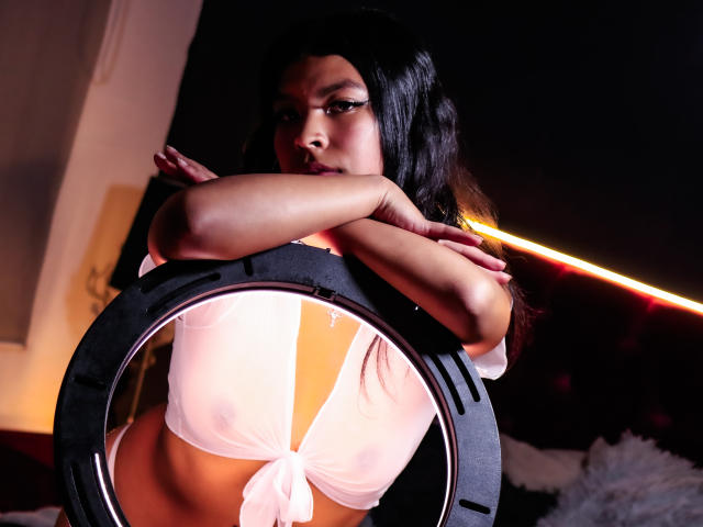 SashaLia with long black hair, arms crossed, wearing a tied-up white crop top, revealing her tits and a neon orange light behind her. Her face is serious, dark background.
