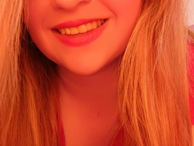 NatalyHamis Gesicht, mit einem lächelnden Mund, rotem Lippenstift, blonden Haaren und einem pinken Oberteil, wobei ihre Zähne und ihre leicht sichtbare Nase hervorgehoben werden..