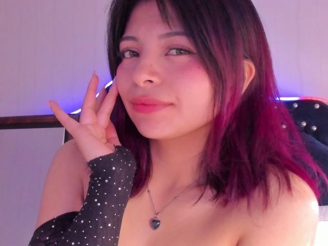ShofiaWett with pinkish-red hair, dark roots, wearing a black polka-dot sleeve, heart necklace, and pink lipstick, smiling softly, hand near face, showing off her tits.