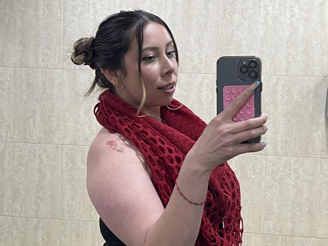 JennyLawry with red leopard print scarf, taking a selfie in a bathroom, showing a bruise on her upper arm, holding a pink phone case, smiling slightly.