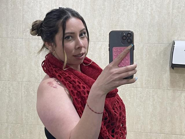 JennyLawry with red scarf, holding phone, looking back at mirror, showing tits in black top, blonde hair in bun, earring, and tattoo on right shoulder. Bathroom setting.
