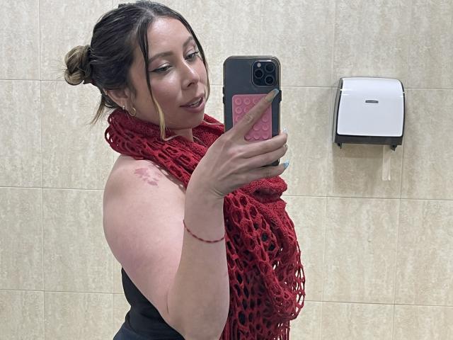 JennyLawry, avec une écharpe en crochet rouge, prend un selfie devant un miroir dans une salle de bain. Elle montre un tatouage sur son épaule et ses seins sont partiellement couverts par un haut noir..