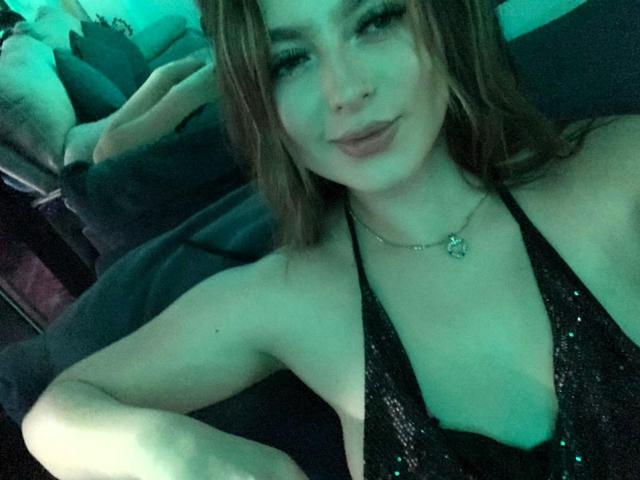 SamanthaRivera with light skin, brown hair, nose ring, and green-tinted selfie, wearing a black sequin halter top, showing her tits, and a heart necklace, lying on a couch.