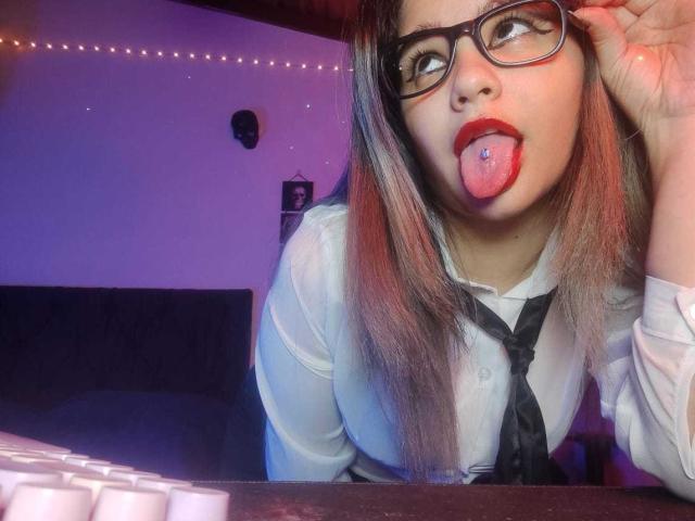 AshGraven with glasses, tongue out, tongue piercing, red lipstick, white shirt, black tie, leaning on desk, looking up, long brown hair, fair skin, slightly bluish lighting.